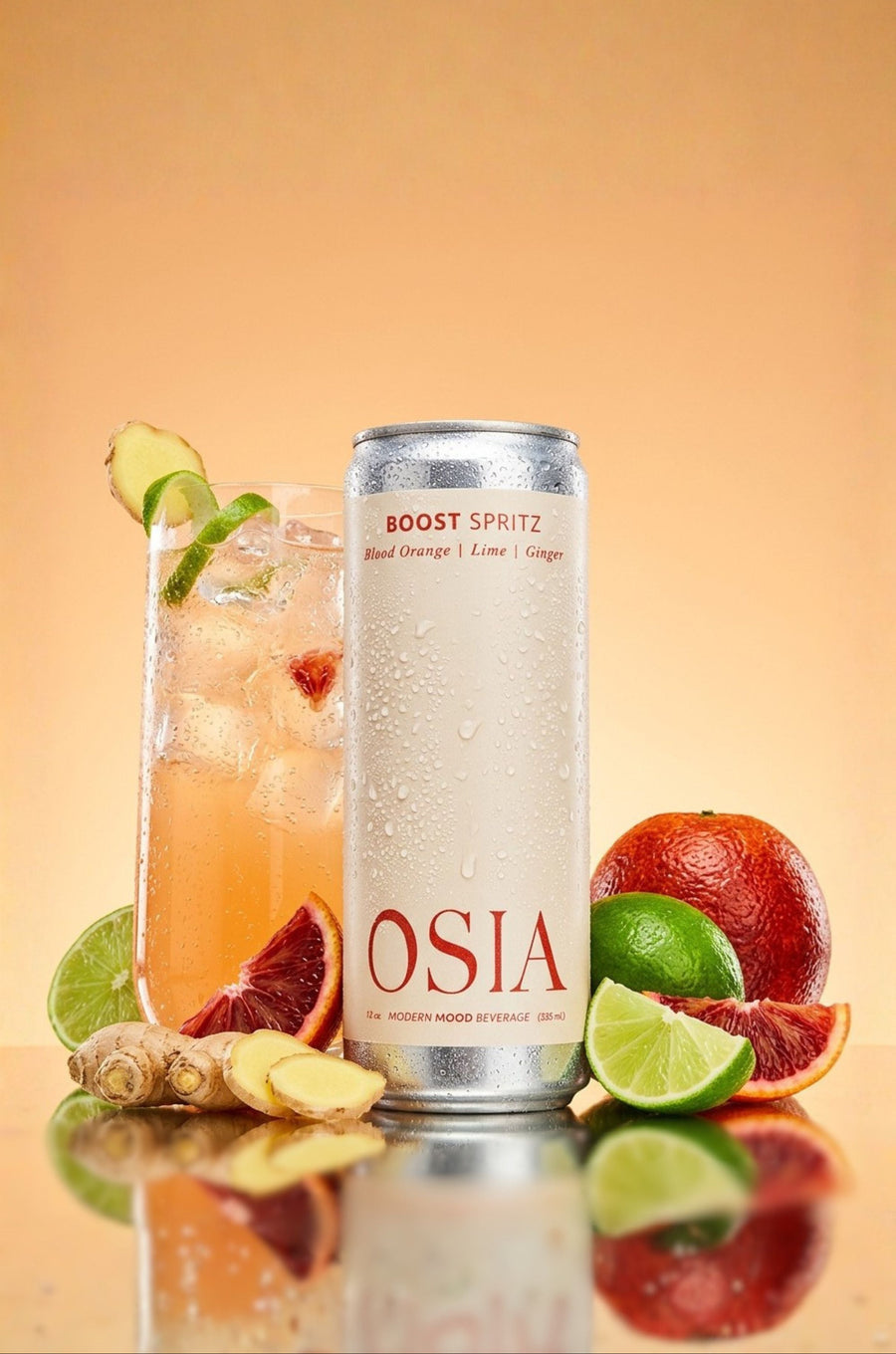 A can of OSIA Boost Spritz beverage stands on a reflective surface beside a tall glass of iced orange drink garnished with lime and ginger, with blood orange slices, lime wedges, and ginger roots against a warm beige gradient background.